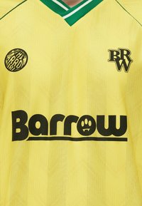 Camiseta deportiva amarilla con ribetes verdes, logo de texto negro "Barrow" con una huella de pata, insignia circular "The Barrow World" y emblema BRW.