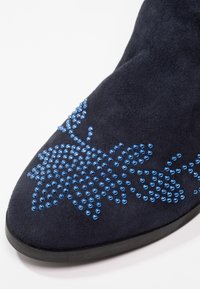 Botín de ante azul oscuro hasta el tobillo con punta afilada, decorado con un patrón floral hecho de pequeñas piedras de strass azules en la parte superior.