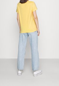 Camiseta de algodón amarilla con mangas cortas, ajuste relajado y dobladillo redondeado. Combinada con jeans de denim azul claro y zapatillas blancas.