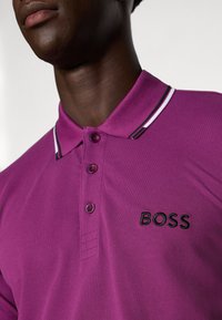 Vijolična polo majica s ovratnikom, ki ima tri gumbke, izvezeno "BOSS" logotip v črni barvi in črtaste poudarke na ovratniku.