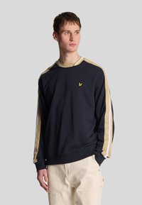 Sweatshirt bleu marine avec des rayures beiges et blanches sur les manches, col rond côtelé et petit logo brodé sur la poitrine. Fabriqué en tissu doux.