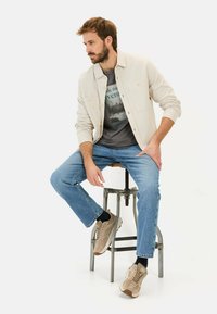 Lys beige skjortejakke over en grå grafisk t-shirt, blå jeans og beige sneakers. Sidder på en metalstol. Bløde teksturer, afslappet design.