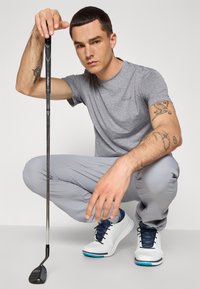 Šedé tričko s krátkým rukávem a světle šedé kalhoty, které má na sobě muž v podřepu, držící golfovou hůl. Bílé boty s modrými akcenty dokončují tento outfit.
