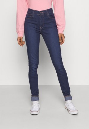 Personne portant un jean skinny bleu foncé avec les revers roulés, un haut à manches longues rose et des baskets blanches, debout sur un sol uni.