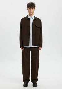 Brun oversized jakke med brystlomme over en blå stripete skjorte, kombinert med løse brune bukser og svarte sko. Enkel, strukturert design.