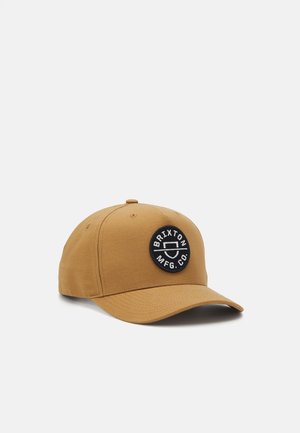 Casquette de baseball marron avec une visière incurvée et un patch circulaire noir portant l'inscription "Brixton MFG. Co." sur le panneau avant.