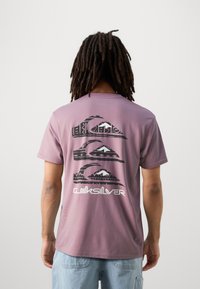 Camiseta de manga corta lavanda con un diseño gráfico de olas y montañas en negro, con el texto "Quiksilver" en la parte inferior.