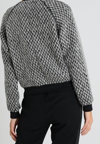 Svetr s šedým a černým houndstooth vzorem, raglánovými rukávy, žebrovanými manžetami a spodním lemem. Měkká textura, mírně volný střih.