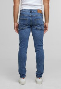 Kék farmernadrág, slim fit szabású, két hátsó zsebbel, barna bőrpánttal a deréknál és struktúrált felülettel. Fehér sportcipőt viselnek.
