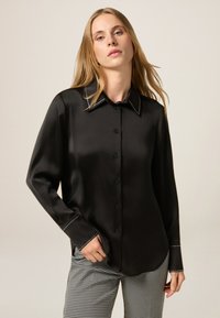 Camicia nera in satin con colletto a punta e cuciture bianche a contrasto. Maniche lunghe, vestibilità comoda e texture liscia.