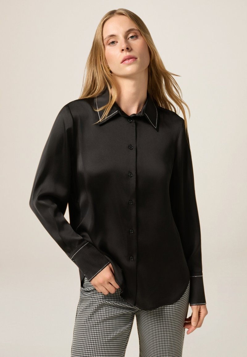 Camicia nera in satin con colletto a punta e cuciture bianche a contrasto. Maniche lunghe, vestibilità comoda e texture liscia.
