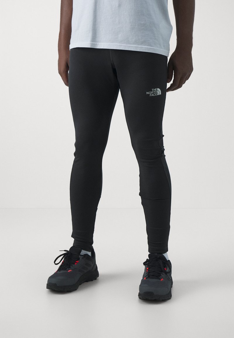 Leggings noirs moulants à la texture lisse, avec un petit logo blanc sur la cuisse, assortis à des chaussures de sport sombres.