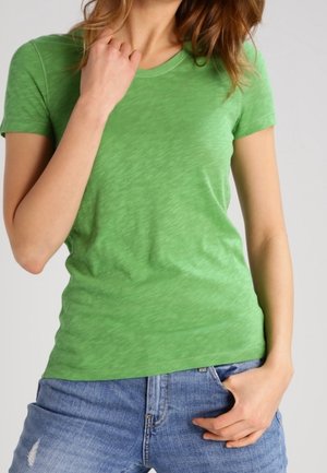 Lyhythihainen vihreä T-paita, joka on valmistettu pehmeästä, hieman teksturoidusta kankaasta. Siinä on pyöreä pääntie, ja se on yhdistetty sinisiin denimhousuihin.