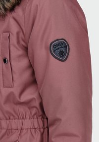 Doudoune bordeaux avec une finition texturée, taille élastique et un écusson noir en forme de bouclier sur la manche gauche portant le nom de la marque "ONLY".