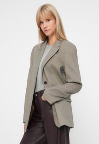 Textúrázott, barna kockás blézer egy gombos záródással, kerekített hajtókákkal és lezser fazonnal, szürke felsővel és sötét nadrággal párosítva.