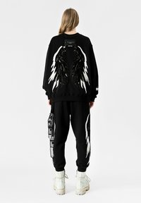 Sudadera negra y pantalones de chándal con gráficos de alas blancas; detalle de lentejuelas texturizadas en la parte posterior. Incluye etiquetas del logo en los pantalones. Botas blancas.