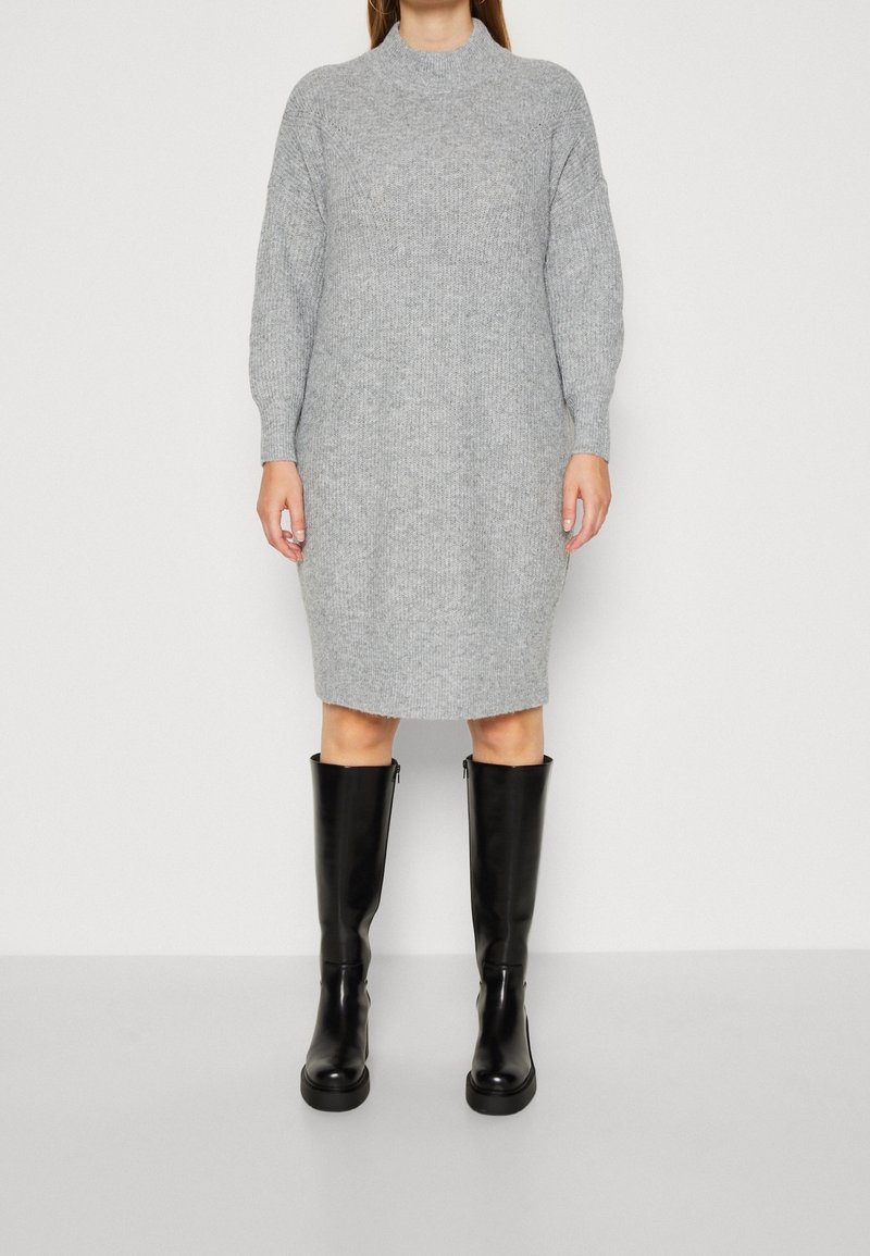 Robe grise en maille avec un col montant et des manches longues, associée à de grandes bottes en cuir noir. La robe a une coupe décontractée et une texture côtelée.