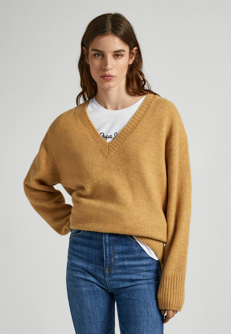 Maglione giallo senape a scollo a V realizzato in materiale morbido, con una vestibilità comoda e maniche lunghe a coste, indossato sopra una maglietta bianca e jeans.