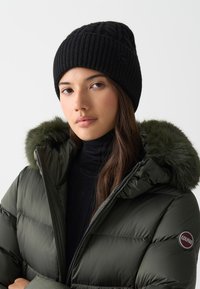 Caciula neagră tricotată cu textură cu rigle și logo; geacă pufă oliv închis cu detalii din blană sintetică la glugă și umplutură cu puf.