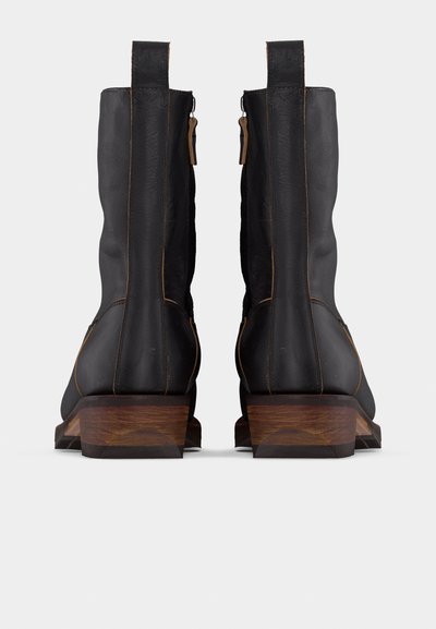 Μαύρες δερμάτινες μπότες μέχρι τη γάμπα με καφέ ξύλινα τακούνια, ορατά φερμουάρ στην πίσω πλευρά και θηλιές για εύκολο φόρεμα, εμφανιζόμενες από πίσω σε λευκό φόντο.