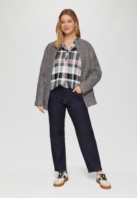 Grijze, gestructureerde buitenjas over een zwart-wit geruite blouse met roze accenten. Donkerblauwe jeans en beige sneakers met zwarte accenten.