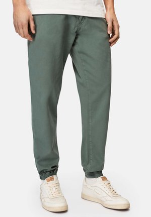 Grüne cropped Joggerhosen mit elastischen Bündchen und Fronttaschen, kombiniert mit weißen Turnschuhen. Das Material hat eine glatte, matte Oberfläche.