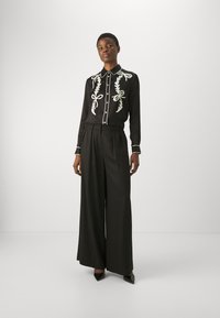 Blusa nera con design floreale ricamato bianco e bottoni, abbinata a pantaloni neri a vita alta e gamba larga e tacchi a punta neri.