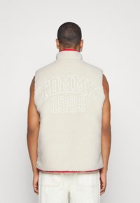 Krémově zbarvená fleecová vesta s vysokým límcem, červeným lemem a vystouplým nápisem "TOMMY 1985" na zádech. Má bezrukávový střih a texturovaný povrch.