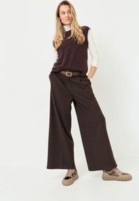 Pantaloni a gamba larga marrone scuro abbinati a un maglione senza maniche viola scuro sopra una camicia bianca a maniche lunghe, completati da sneakers beige con motivo.