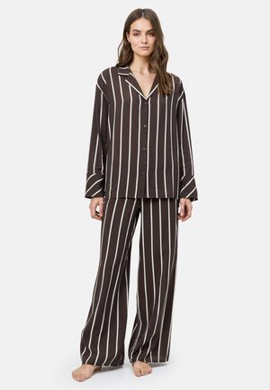 Vrouw staat blootsvoets en draagt losse donkerbruine pyjama met verticale witte strepen, een hemd met knopen en broek met wijde pijpen.