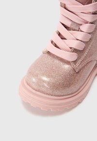 Rosa glittrande ankelstövlett med slät, glänsande yta, rund tå och texturerad gummisula. Snören av ljusrosa vävt tyg.