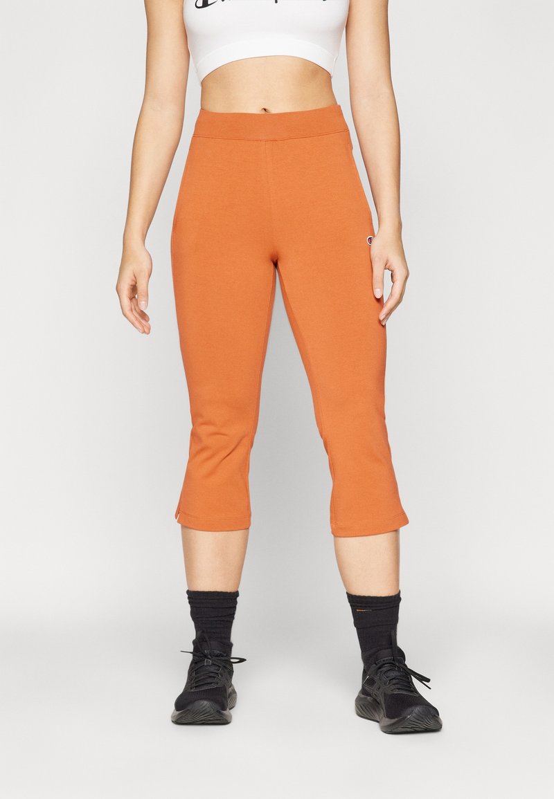 Femme portant un pantalon court orange, des chaussettes noires, des baskets noires et un haut court blanc sur un fond clair uni.
