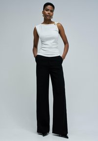 Top sin mangas blanco texturizado con un hombro y detalle en oro, combinado con pantalones de pierna ancha negros de cintura alta y tacones puntiagudos.