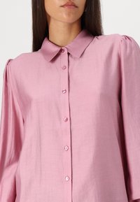 Camisa botão rosa claro com colarinho, apresentando mangas longas com detalhes suaves de volume e uma textura de tecido suave e macia.