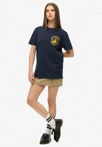 Camiseta de algodón azul marino con una impresión gráfica amarilla en el lado izquierdo del pecho, combinada con una falda caqui y zapatos negros, que lleva calcetines a rayas blancas y negras.