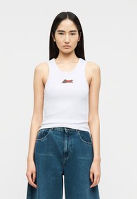 Νεαρή γυναίκα με μακριά σκούρα μαλλιά, που φοράει ένα λευκό αμάνικο με μικρό σχέδιο πορτοκαλί ψαριού και φαρδιά μπλε τζιν.