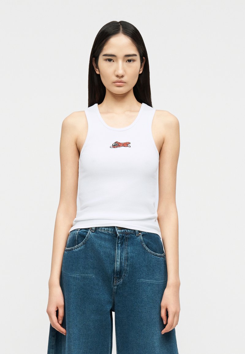 Νεαρή γυναίκα με μακριά σκούρα μαλλιά, που φοράει ένα λευκό αμάνικο με μικρό σχέδιο πορτοκαλί ψαριού και φαρδιά μπλε τζιν.