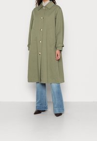 Olijfgroene katoenen trenchcoat met een brede kraag, knopen aan de voorkant en zijzakken. Gepaard met wijd uitlopende spijkerbroeken en schoenen met een puntige neus.