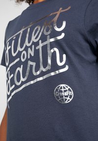 Tmavě modré tričko z bavlny s nápisem ve stříbrné metalické barvě "Nejlepší na Zemi" a kruhovým logem CrossFit.