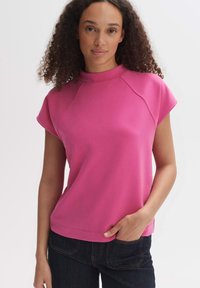 Roze top met korte mouwen en een hoge hals, gemaakt van gladde stof. Kenmerkt zich door raglan mouwen en een relaxed fit, gecombineerd met donkerblauwe jeans.