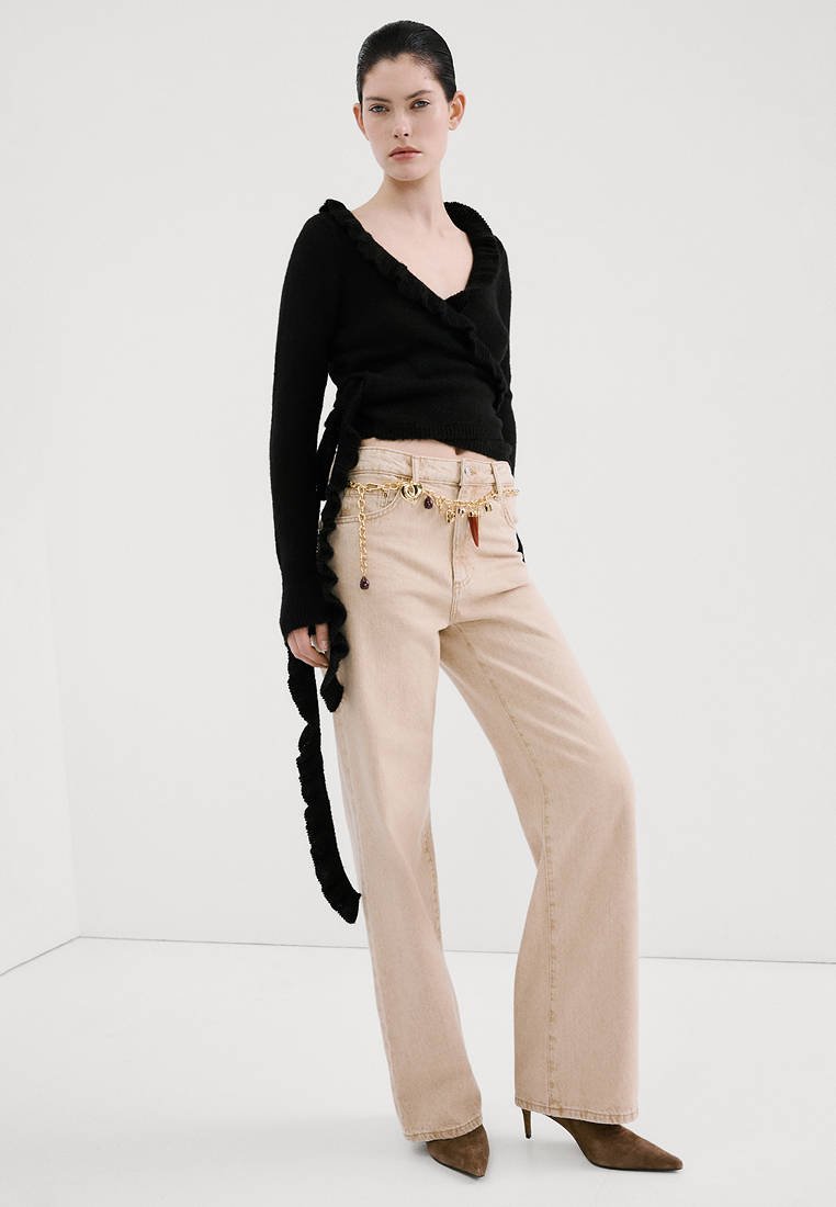 Femme portant un pull noir croisé, un jean beige à jambes larges avec ceinture chaîne dorée et des talons pointus marron, debout devant un fond blanc uni.
