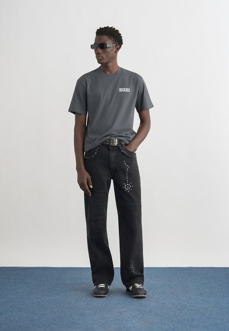 Hombre con gafas de sol oscuras, camiseta gris de Dickies, jeans negros con diseño de tachuelas, cinturón negro y zapatillas negras, de pie sobre una alfombra azul.