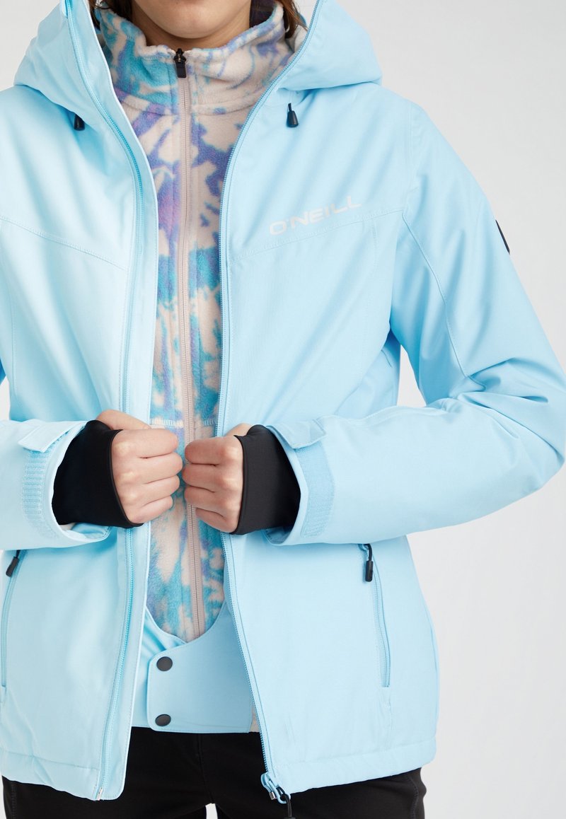 Giacca da sci azzurro chiaro con un design aderente. Presenta un tessuto impermeabile, polsini neri e uno strato interno in fleece a motivi. Dettaglio della zip e del logo visibili.