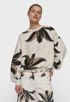 Vrouw draagt een beige sweatshirt en broek met grote zwarte en bruine bloemenprints, en een lichte riem met metalen studs.