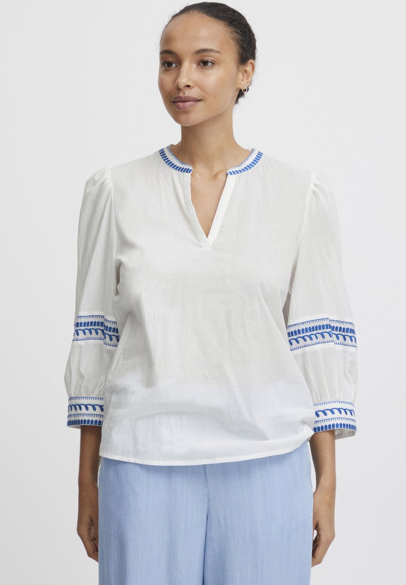 Blouse blanche avec des accents brodés bleus sur les manches et le col, offrant une coupe décontractée et un col en V. Tissu léger.