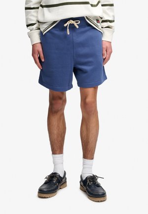 Homme portant un short bleu avec cordon, des chaussettes blanches, des chaussures bateau bleu marine et une chemise blanche à manches longues avec des rayures vertes.