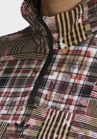 Monivärinen puolivetoketjullinen neule, jossa on ruudullinen kuvio punaisen, mustan ja kullan sävyissä, kudottu tekstuuri ja pieni logo helmassa.