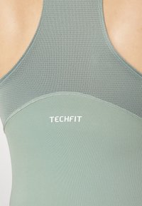 Zaļš sporta topiņš ar sieta tekstūru, ar gludu, pieguļošu dizainu. Uzmanību pievērsto "TECHFIT" logo.
