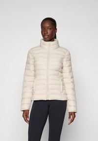 Pufferjack in licht crèmekleur met een boxy ontwerp, hoge kraag, rits aan de voorkant en een textuur gequilt patroon. Draagt over een donkere legging.