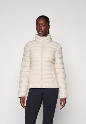 Puffjacka i ljus krämfärg med fyrkantig design, hög krage, framdragkedja och texturerad quiltad mönstring. Bärs över mörka leggings.